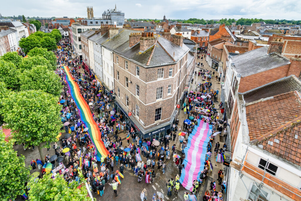 Aerial view of York Pride parade with vibrant rainbow and pink, blue, white flags lining the street, full of attendees.