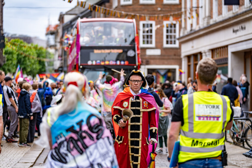 City crier in vibrant Pride attire leads York Pride parade, surrounded by supportive crowd and rainbow flags.