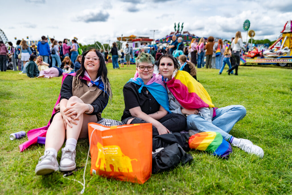 Three people sit on the grass wearing pride flags at York Pride, smiling and enjoying the festive atmosphere.