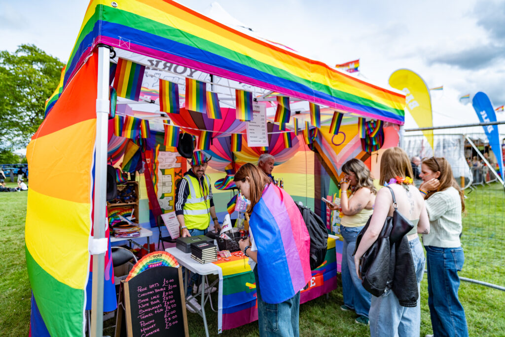 Pride stall with rainbow flags and visitors exploring colourful merchandise at York Pride festival.
