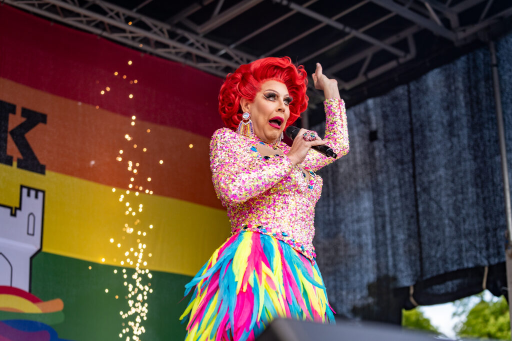 Drag performer in sparkly outfit and rainbow feathers entertains on stage at York Pride with lively expression.