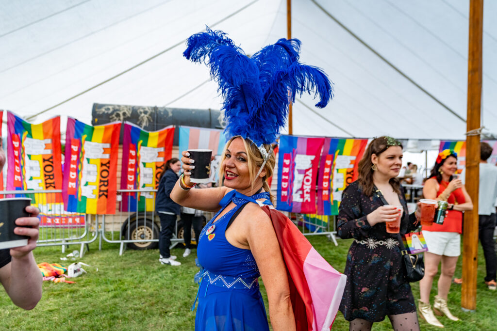 Person in vibrant blue attire and feathered hat enjoys York Pride festival, holding a drink with colourful banners behind.