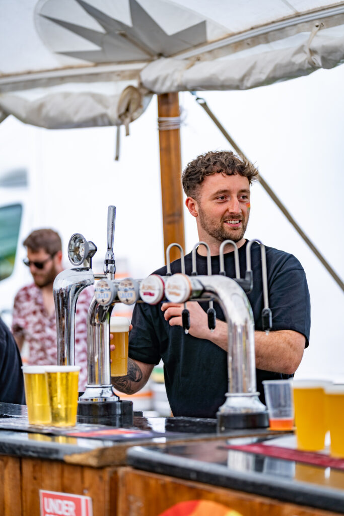 Bartender with rainbow face paint smiling at York Pride event outdoor bar, serving drinks to attendees.