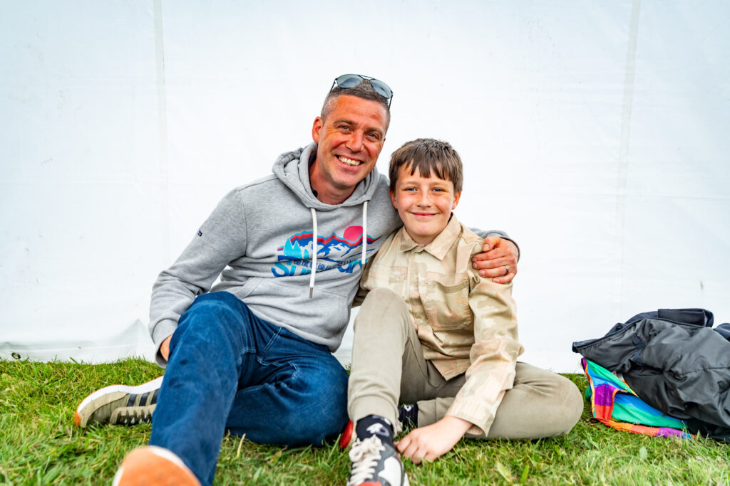 Two people smiling and sitting on grass at York Pride, with a rainbow flag nearby. Happy, inclusive atmosphere.