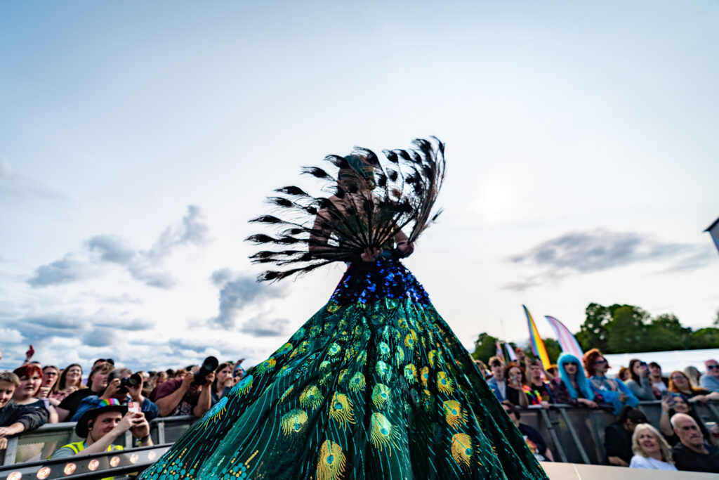 Performer in peacock-inspired costume on stage at York Pride, with crowd and flags in the background.