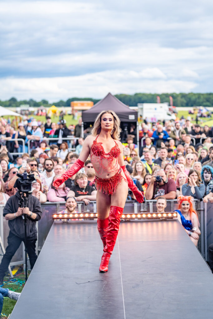 Drag performer in red costume struts on stage at York Pride, with a lively crowd in the background.